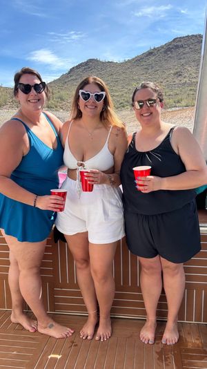 Three friends on a boat at a desert lake, wearing swimsuits and sunglasses and holding red cups, smiling in front of rocky, cactus-covered hills under a bright blue sky.