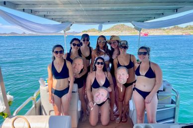 Group of women in black bikinis posing and laughing on a covered pontoon boat over turquoise coastal waters, holding novelty face cutouts