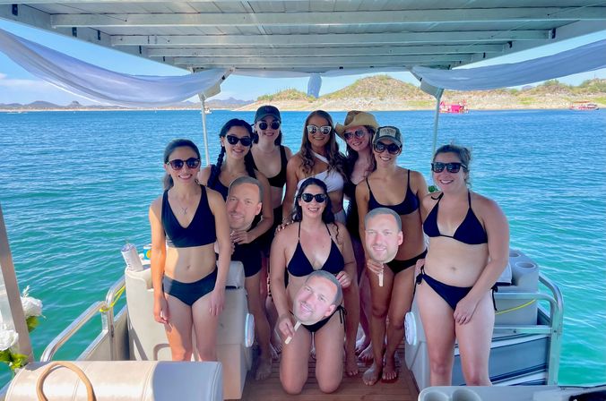 Group of women in black bikinis posing and laughing on a covered pontoon boat over turquoise coastal waters, holding novelty face cutouts