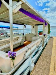 Pontoon party boat docked at a lakeside pier with cream lounge seating, purple drapes and floral accents under a bright blue sky and distant hills.