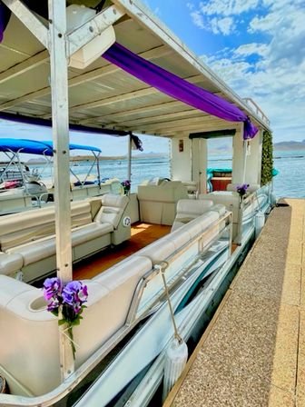 Pontoon party boat docked at a lakeside pier with cream lounge seating, purple drapes and floral accents under a bright blue sky and distant hills.