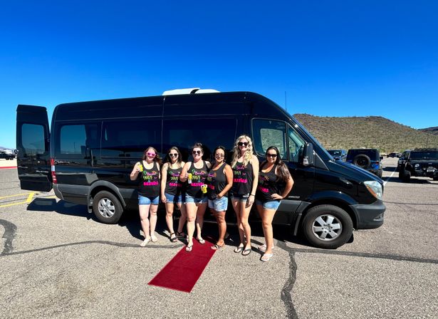 Six women in matching neon tank tops and denim shorts holding drinks and posing on a short red carpet beside a black party van in a sunny desert parking lot with clear blue sky and low scrubby hills.