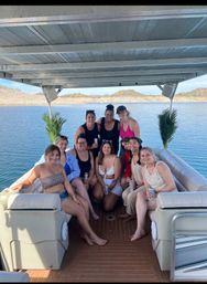 Group of friends enjoying a sunny lake day on a covered pontoon boat, smiling and holding canned drinks with rocky, arid hills in the background