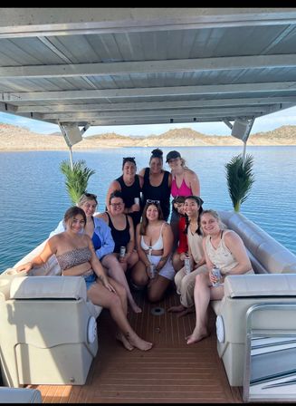 Group of friends enjoying a sunny lake day on a covered pontoon boat, smiling and holding canned drinks with rocky, arid hills in the background
