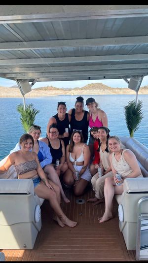 Group of friends enjoying a sunny lake day on a covered pontoon boat, smiling and holding canned drinks with rocky, arid hills in the background