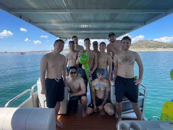 Smiling group of shirtless friends posing with a green inflatable frog on a covered pontoon boat over a sunny blue lake with rocky shoreline and clear sky.