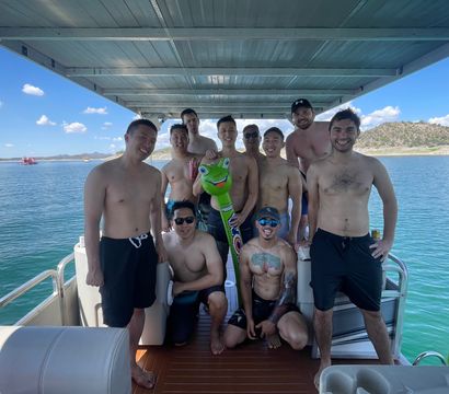 Smiling group of shirtless friends posing with a green inflatable frog on a covered pontoon boat over a sunny blue lake with rocky shoreline and clear sky.
