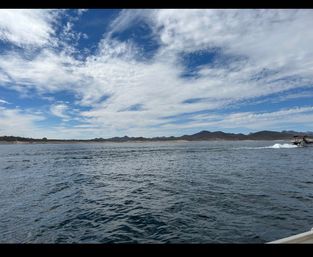 Wide lake scene with rippling blue water, low mountains along the shoreline, a dramatic wispy-cloud blue sky, and a small motorboat leaving a white wake at the right edge.