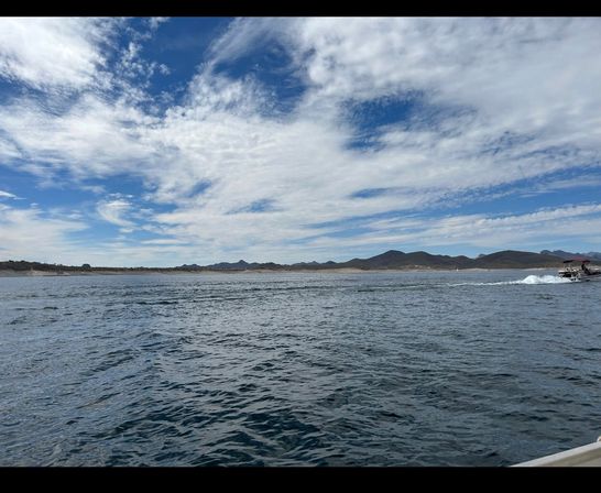 Wide lake scene with rippling blue water, low mountains along the shoreline, a dramatic wispy-cloud blue sky, and a small motorboat leaving a white wake at the right edge.