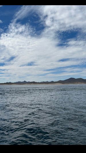 Wide lake scene with rippling blue water, low mountains along the shoreline, a dramatic wispy-cloud blue sky, and a small motorboat leaving a white wake at the right edge.