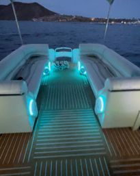 Boat deck at dusk with glowing aqua LED lights illuminating cushioned bench seating and teak-style flooring, calm sea and distant shoreline.