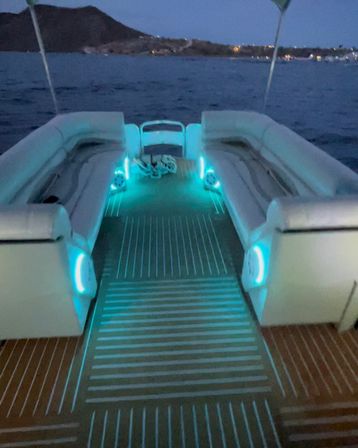Boat deck at dusk with glowing aqua LED lights illuminating cushioned bench seating and teak-style flooring, calm sea and distant shoreline.