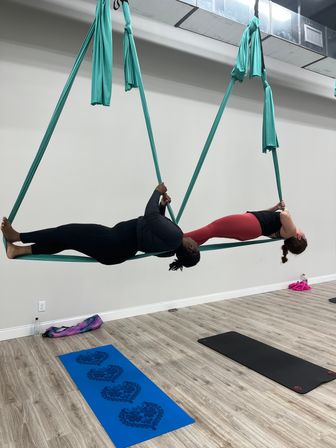 Two women practicing aerial yoga on teal silk hammocks, suspended horizontally in a bright indoor studio above wood flooring with blue and black yoga mats below.