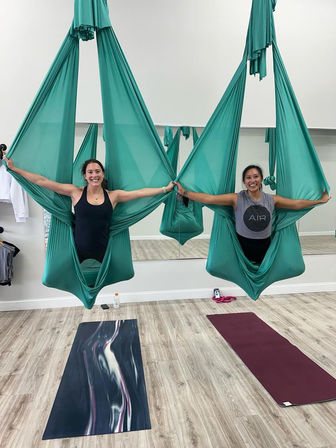 Two smiling women suspended in teal aerial yoga hammocks, holding hands in a bright studio above yoga mats — aerial yoga class scene