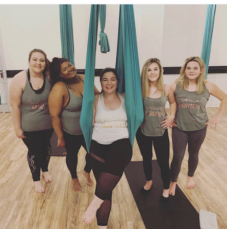 Five women smiling in a group aerial yoga class in a bright studio with teal hammocks and wood flooring