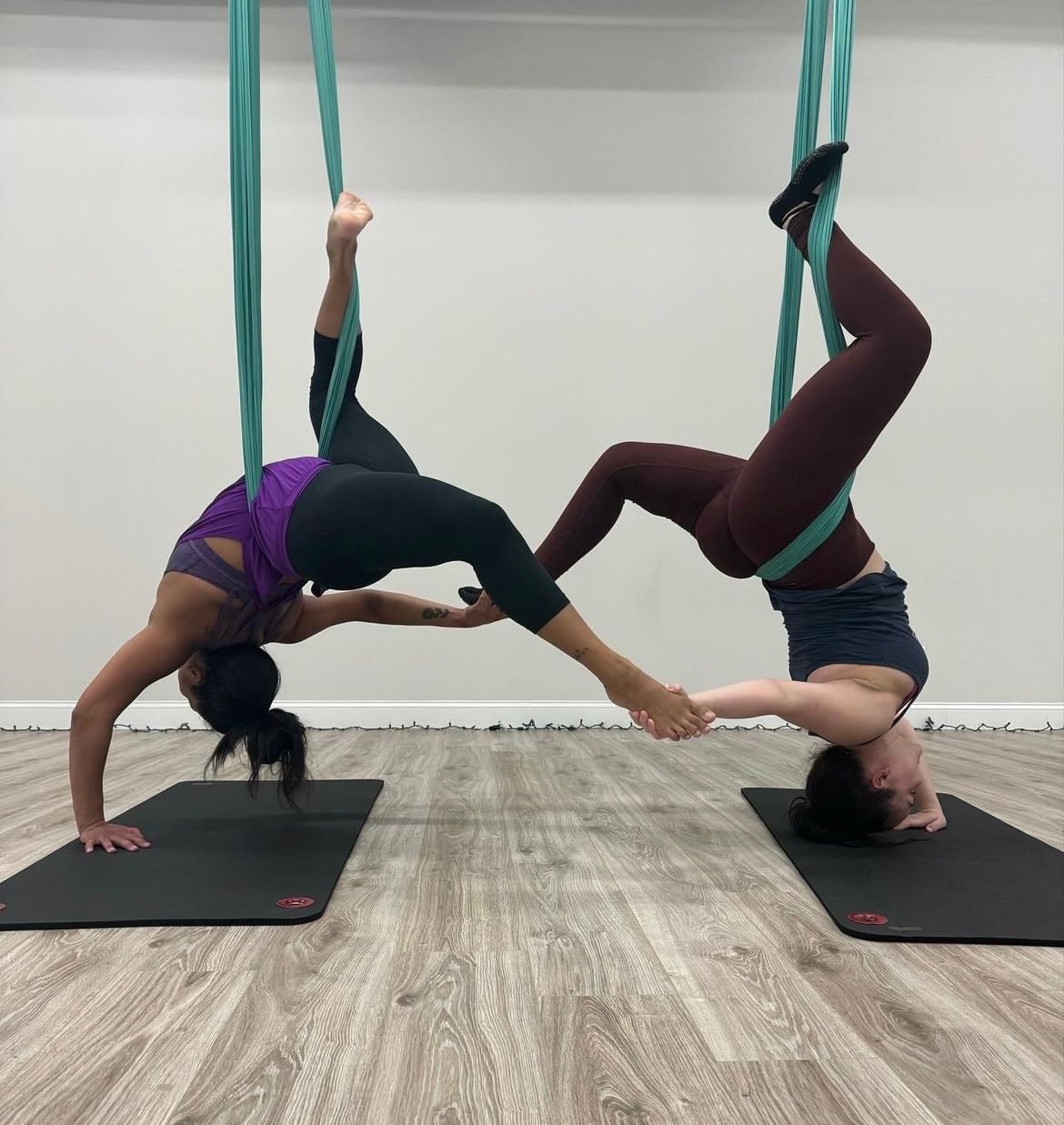 Two people in an indoor aerial yoga studio suspended in teal silks, performing mirrored inverted poses and holding each other’s feet above wood floors and yoga mats.