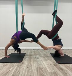 Two people in an indoor aerial yoga studio suspended in teal silks, performing mirrored inverted poses and holding each other’s feet above wood floors and yoga mats.