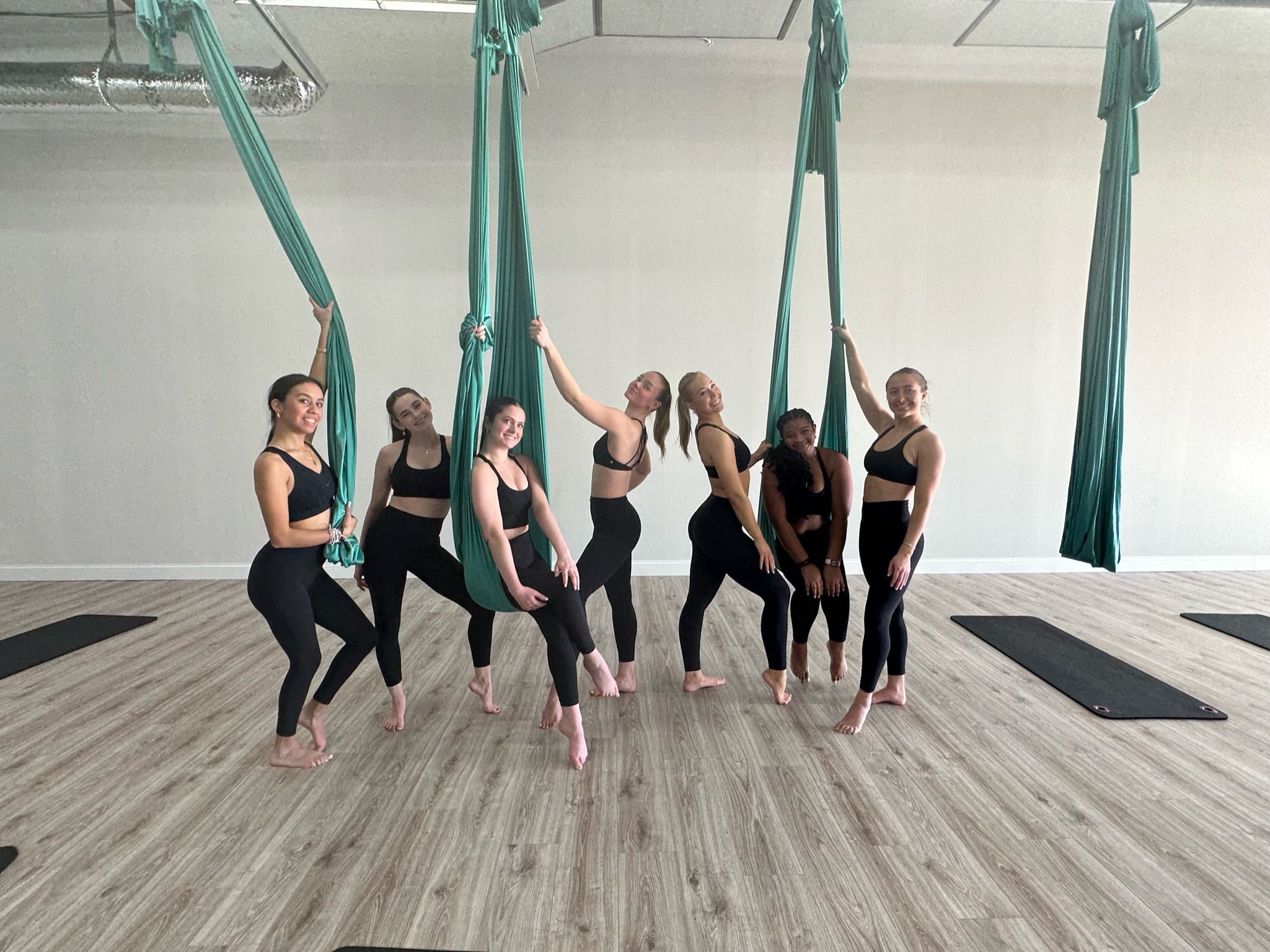 Group of seven women in black activewear striking playful poses with teal aerial yoga hammocks in a bright studio with wood-look floors and yoga mats