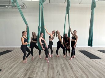 Group of seven women in black activewear striking playful poses with teal aerial yoga hammocks in a bright studio with wood-look floors and yoga mats
