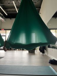 Teal aerial yoga hammock suspended in a bright studio over a yoga mat on light wood flooring, an arm peeking from inside.
