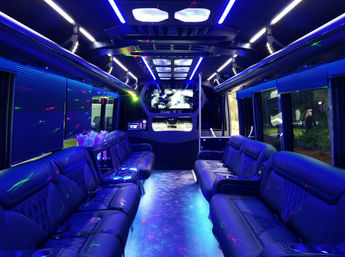 Luxury party bus interior with blue LED ambient lighting, plush black leather lounge seating along both sides, center aisle, mounted flat-screen TV, stainless dance pole, cupholders and multicolored laser accents — perfect for urban nightlife and event transportation.
