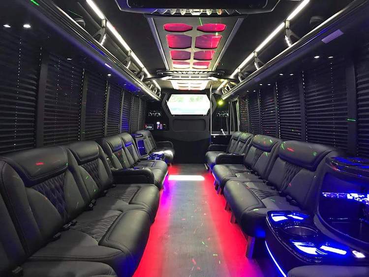 Luxury party bus interior with black leather bench seats along both sides, red and purple LED floor and ceiling lights, cup holders and dark window shades for a nightlife or event ride.