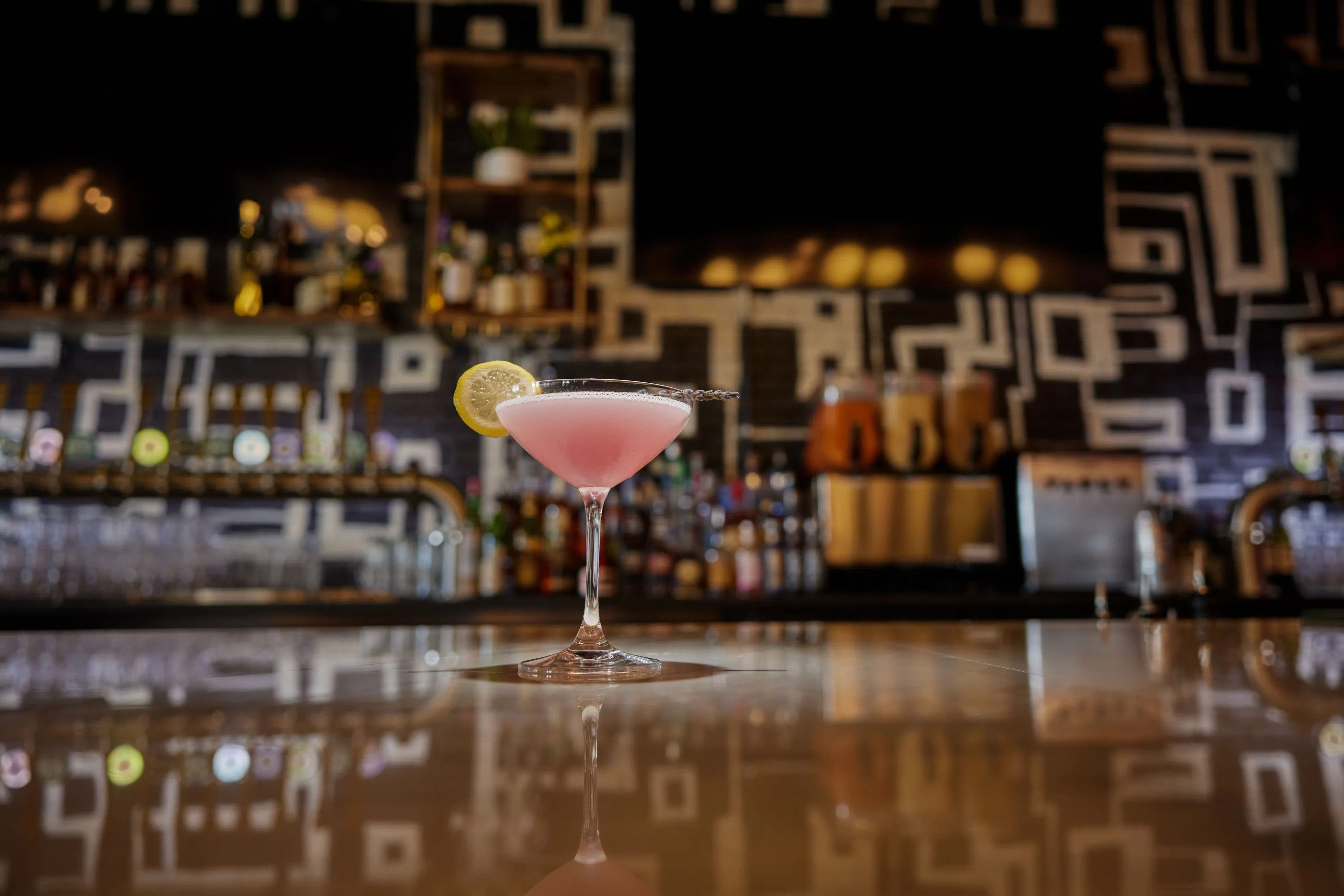 Cheery pink cocktail in a martini glass with a lemon wheel, sitting on a glossy bar counter reflecting a modern geometric-tiled back bar and draft taps