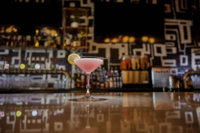 Cheery pink cocktail in a martini glass with a lemon wheel, sitting on a glossy bar counter reflecting a modern geometric-tiled back bar and draft taps