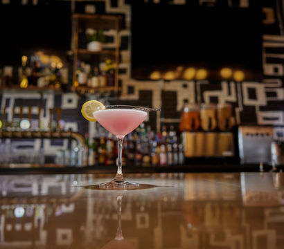 Cheery pink cocktail in a martini glass with a lemon wheel, sitting on a glossy bar counter reflecting a modern geometric-tiled back bar and draft taps