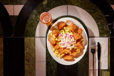 Top-down view of creamy mac and cheese topped with crispy fried chicken bites and colorful coleslaw in a white bowl, served with a small glass of beer and fork and knife on a green-and-white marble restaurant table.
