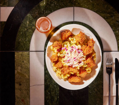 Top-down view of creamy mac and cheese topped with crispy fried chicken bites and colorful coleslaw in a white bowl, served with a small glass of beer and fork and knife on a green-and-white marble restaurant table.