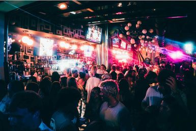 Packed neon-lit bar with crowds at a backlit counter, globe chandeliers and colorful mural — lively downtown nightlife scene.