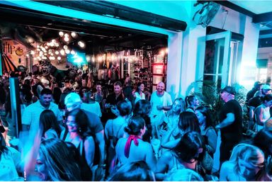Busy indoor-outdoor bar scene at night: crowd of people mingling with drinks under blue neon and globe string lights, mural art and lively patio vibes.