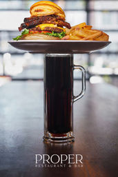 Stacked bacon cheeseburger with melted cheese, lettuce and tomato and thick-cut fries on a plate perched atop a tall glass of cola on a wooden restaurant table with a blurred background.