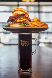Towering gourmet cheeseburger with melted cheese, bacon, crispy onion rings, lettuce and tomato served with thick-cut fries on a white plate balanced atop a tall mug of dark craft beer in a bright pub setting.