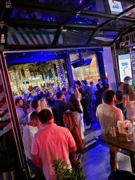 Lively nighttime outdoor bar patio with people mingling under blue-purple lights, open garage-style door, palm trees wrapped in string lights and an illuminated downtown street beyond.