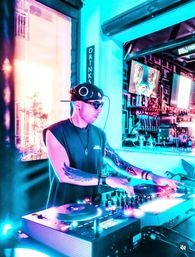 Neon-lit DJ at a nightclub bar mixing on turntables in sunglasses and headphones, tattooed arms visible, vibrant cyan and magenta lighting with drink taps and neon artwork in the background.