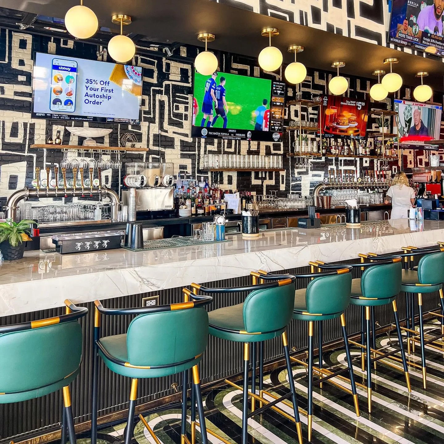 Vibrant urban bar interior with marble countertop, emerald leather stools with brass trim, globe pendant lights, wall TVs showing sports, chrome beer taps and geometric black-and-white mural.