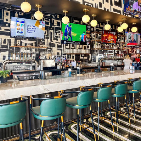 Vibrant urban bar interior with marble countertop, emerald leather stools with brass trim, globe pendant lights, wall TVs showing sports, chrome beer taps and geometric black-and-white mural.