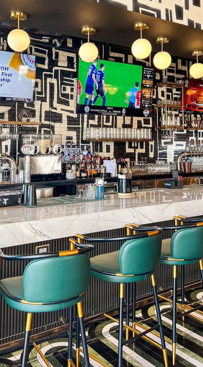 Vibrant urban bar interior with marble countertop, emerald leather stools with brass trim, globe pendant lights, wall TVs showing sports, chrome beer taps and geometric black-and-white mural.