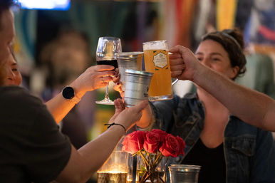 Group of friends toasting with a beer mug, wine glass and metal cups over a table with red roses and candlelight at a cozy bar.