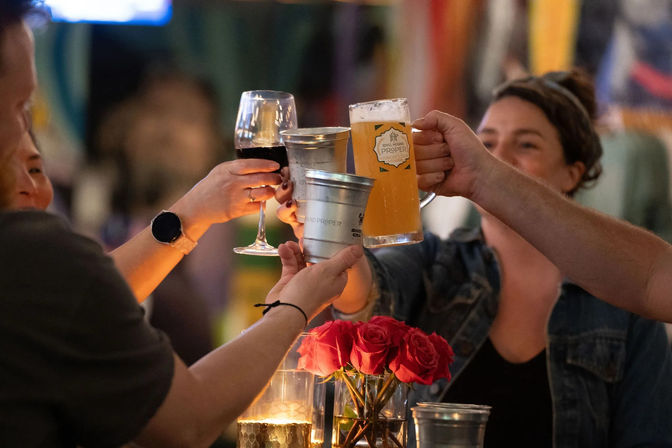 Group of friends toasting with a beer mug, wine glass and metal cups over a table with red roses and candlelight at a cozy bar.