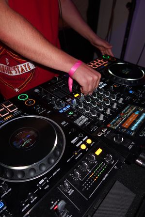Close-up of DJ hands with a pink wristband adjusting knobs on a professional mixer and dual turntables, glowing pads and waveform displays in a dim club booth.