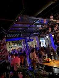 Busy nighttime bar patio with open garage-style door to a tree-lined street, illuminated palm trees and string lights; crowd of people socializing under blue neon lights, wooden communal table with drinks and condiments in the foreground — lively downtown nightlife scene.
