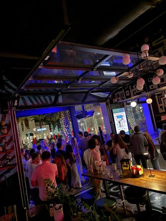 Busy nighttime bar patio with open garage-style door to a tree-lined street, illuminated palm trees and string lights; crowd of people socializing under blue neon lights, wooden communal table with drinks and condiments in the foreground — lively downtown nightlife scene.