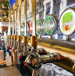 Close-up of colorful craft beer tap handles lined up on a brass draft tower, chrome faucets and round brewery logos (including a crab-themed pilsner label) with glassware underneath in a lively bar setting.