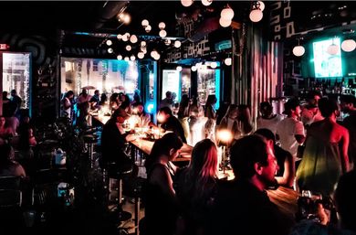 Bustling downtown cocktail bar at night with city nightlife vibe — groups chatting at long wooden communal tables, hanging globe lights, warm table lamps and neon-blue accents.