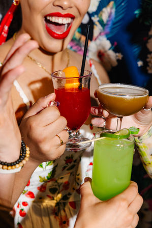 Close-up of friends toasting colorful cocktails at a tropical-themed party — bright red citrus cocktail with orange peel and straw, frothy brown coupe, and neon green lime cocktail, with a joyful red-lipsticked smile in the background.