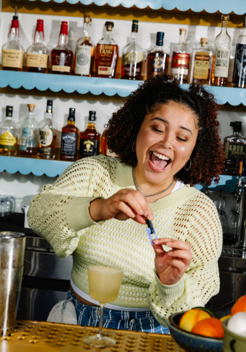 Smiling bartender joyfully adding a garnish to a frothy craft cocktail at a colorful bar counter with shelves of liquor bottles and a bowl of citrus