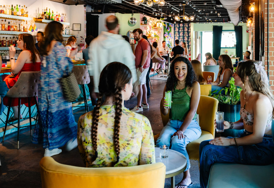 Vibrant neighborhood cocktail lounge with friends laughing over colorful drinks, yellow lounge chairs and tufted bar stools, stocked bar and floral decor in a bustling indoor setting